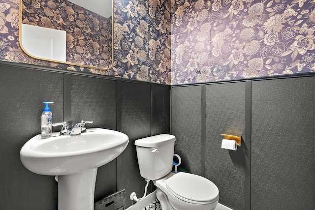 half bathroom with a decorative wall, wainscoting, toilet, and wallpapered walls