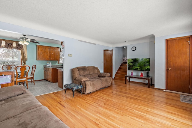 living room featuring light wood finished floors, baseboards, ceiling fan, stairs, and a textured ceiling