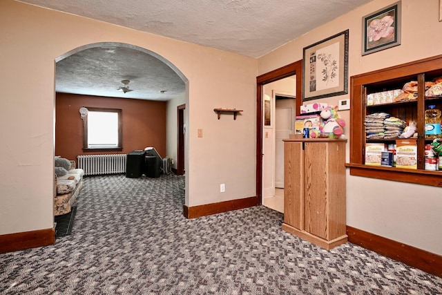 corridor featuring arched walkways, a textured ceiling, baseboards, dark colored carpet, and radiator heating unit