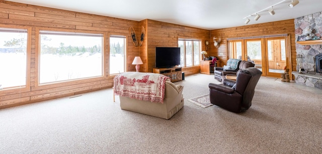 carpeted living area featuring a stone fireplace, track lighting, plenty of natural light, and wooden walls