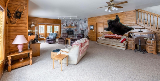 living area featuring stairway, a ceiling fan, carpet flooring, wood walls, and a stone fireplace