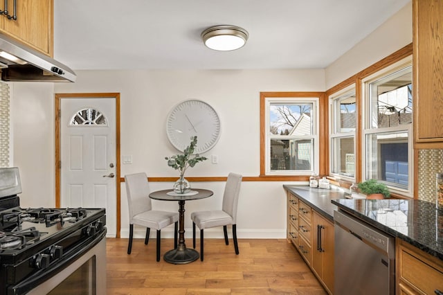 kitchen with dark stone countertops, baseboards, under cabinet range hood, appliances with stainless steel finishes, and light wood-type flooring