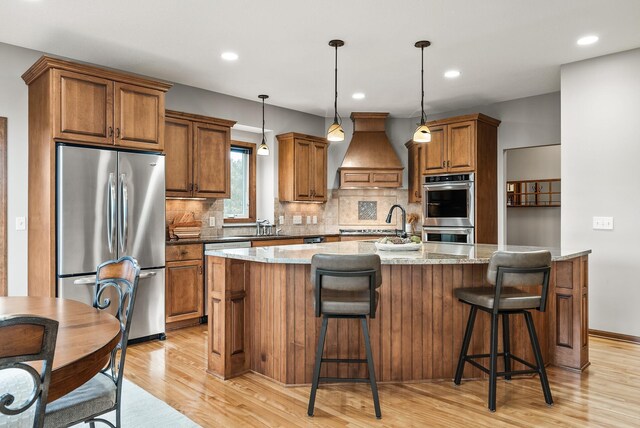 kitchen featuring a center island with sink, brown cabinetry, appliances with stainless steel finishes, custom exhaust hood, and light wood-style floors