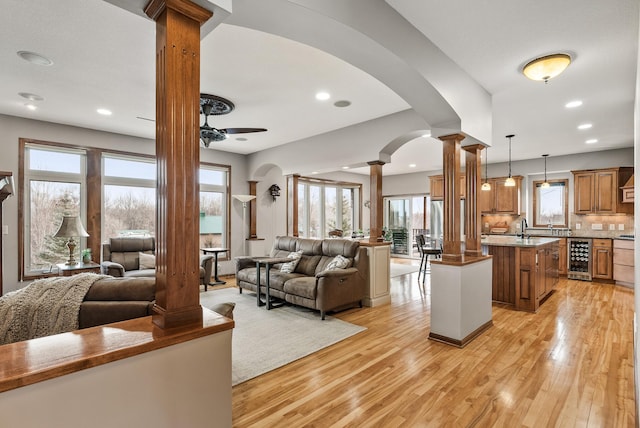 living area featuring beverage cooler, ceiling fan, light wood-style floors, and ornate columns