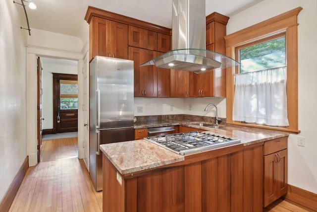 kitchen with island exhaust hood, appliances with stainless steel finishes, brown cabinetry, a sink, and light stone countertops