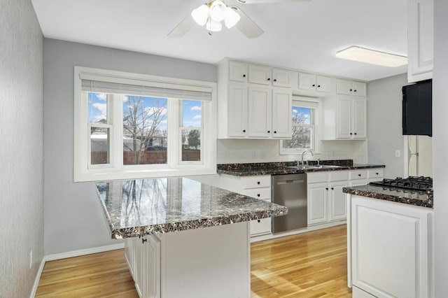 kitchen with a sink, light wood-style floors, white cabinets, stainless steel dishwasher, and dark stone countertops