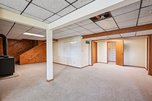 finished below grade area with a paneled ceiling, wooden walls, carpet flooring, baseboards, and a wood stove