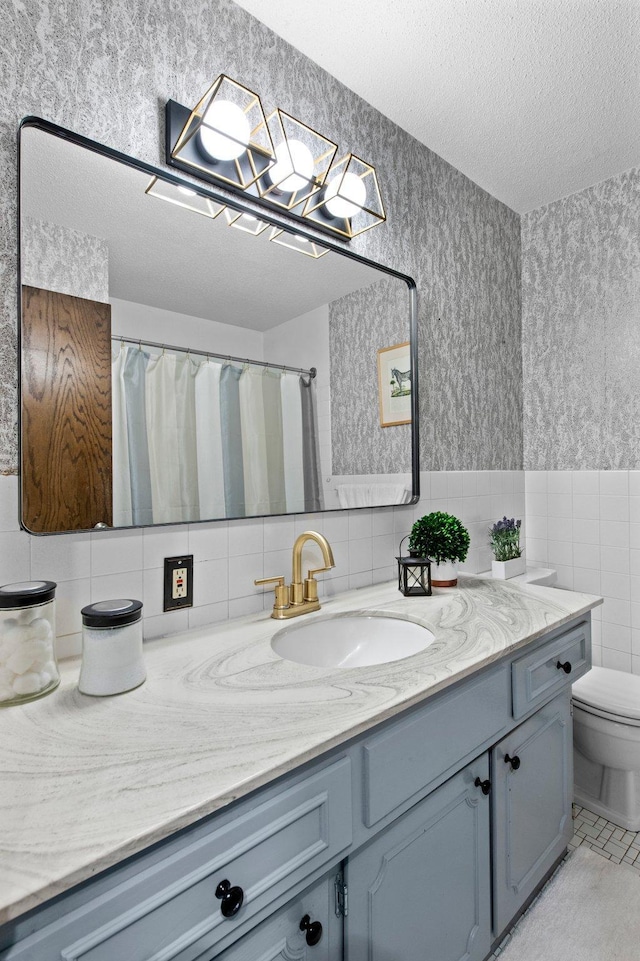 bathroom featuring a textured ceiling, tile walls, vanity, toilet, and wallpapered walls