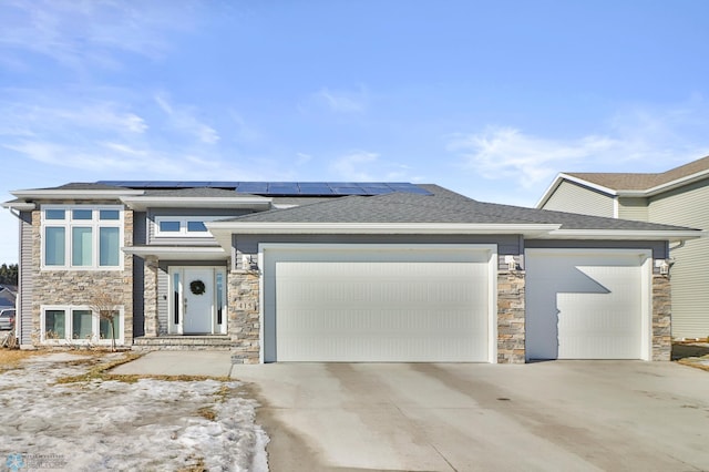 prairie-style home with a garage, driveway, a shingled roof, stone siding, and roof mounted solar panels