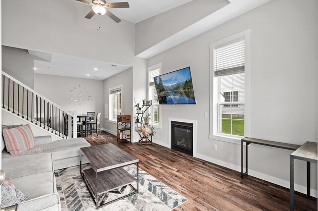 living area featuring stairway, a glass covered fireplace, ceiling fan, wood finished floors, and baseboards