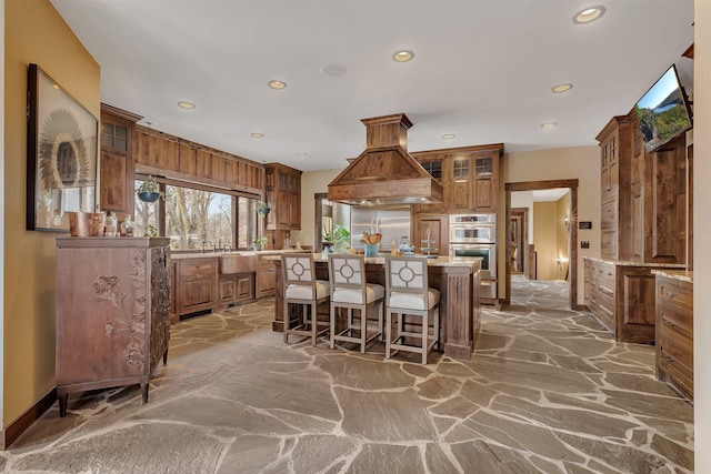 kitchen featuring double oven, island range hood, brown cabinets, and stone flooring