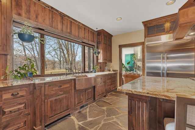 kitchen with glass insert cabinets, light stone countertops, stone finish flooring, a sink, and built in fridge