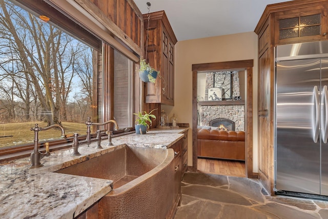 kitchen with light stone counters, stainless steel built in fridge, a fireplace, brown cabinets, and glass insert cabinets