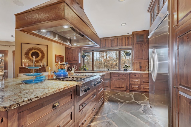 kitchen featuring light stone counters, stainless steel appliances, custom exhaust hood, brown cabinets, and stone finish floor