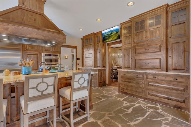 kitchen with custom range hood, light stone counters, brown cabinets, and stone flooring