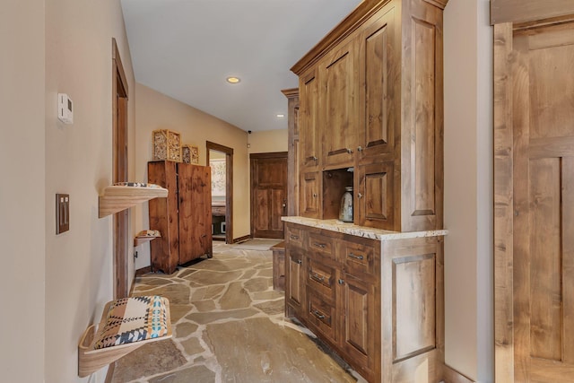 kitchen featuring baseboards, brown cabinetry, light stone countertops, stone floors, and recessed lighting