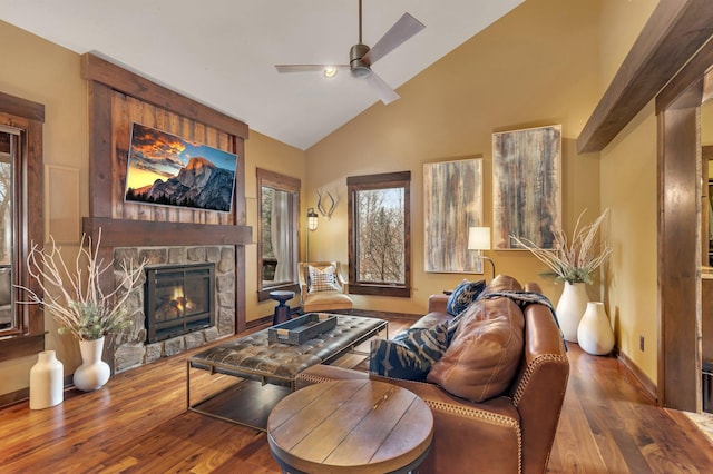 living area with ceiling fan, high vaulted ceiling, a stone fireplace, wood finished floors, and baseboards