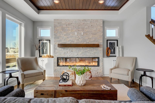 living area with plenty of natural light, a stone fireplace, wooden ceiling, and a raised ceiling