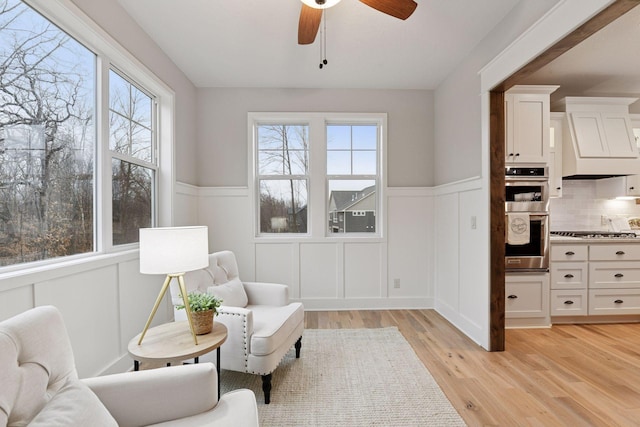 sitting room with light wood-style floors, wainscoting, a decorative wall, and a ceiling fan