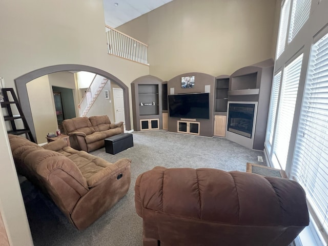 living room featuring a towering ceiling, a glass covered fireplace, stairway, and carpet