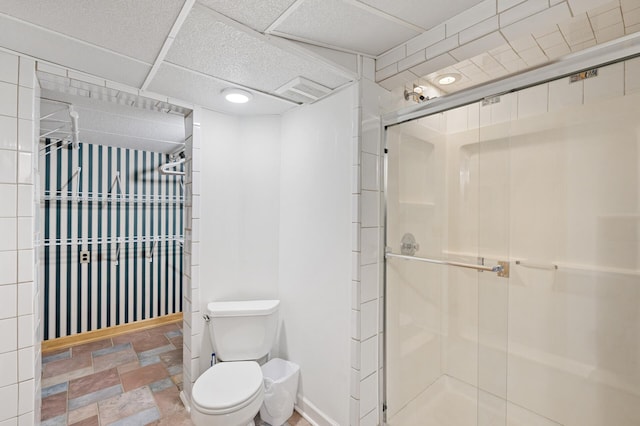 bathroom featuring toilet, a stall shower, a drop ceiling, and stone tile flooring