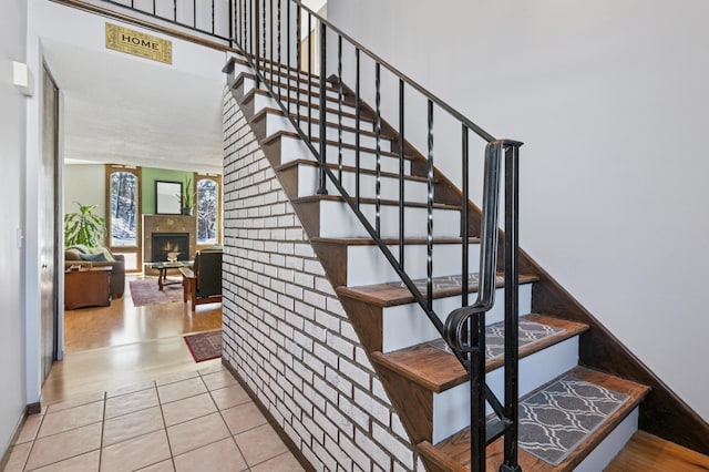 stairs featuring tile patterned floors, brick wall, a towering ceiling, and a lit fireplace