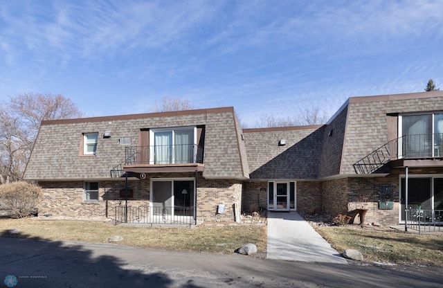 view of front of home featuring brick siding, a balcony, mansard roof, and roof with shingles