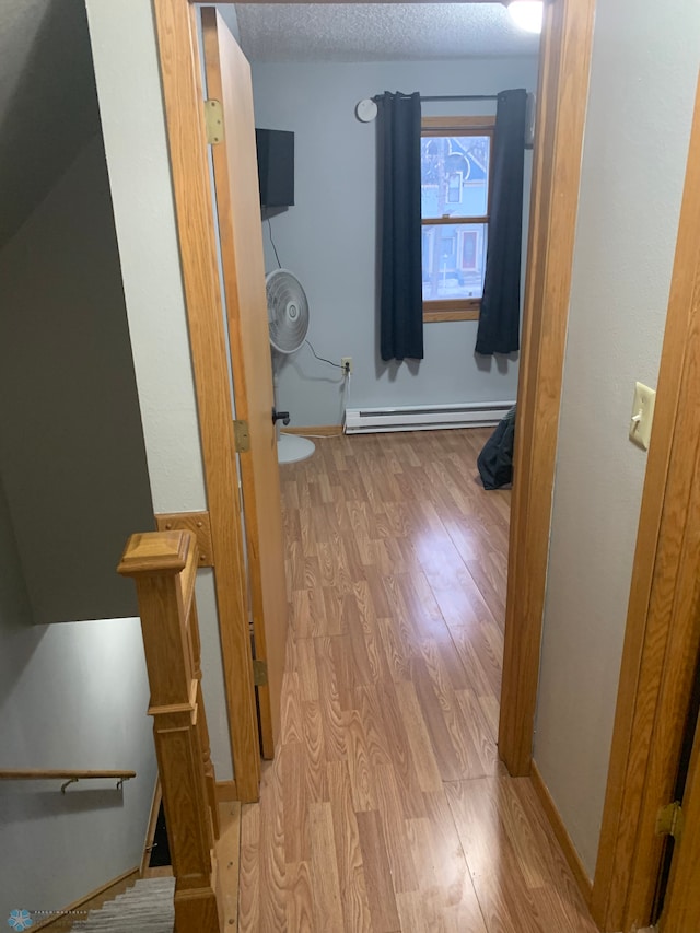 corridor with a textured ceiling, a baseboard heating unit, wood finished floors, an upstairs landing, and baseboards