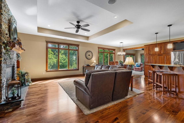 living area featuring a stone fireplace, ceiling fan with notable chandelier, dark wood-style flooring, baseboards, and a raised ceiling