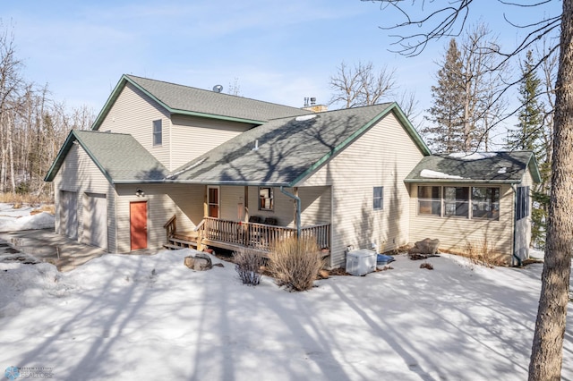 snow covered rear of property featuring a deck, an attached garage, driveway, roof with shingles, and a chimney