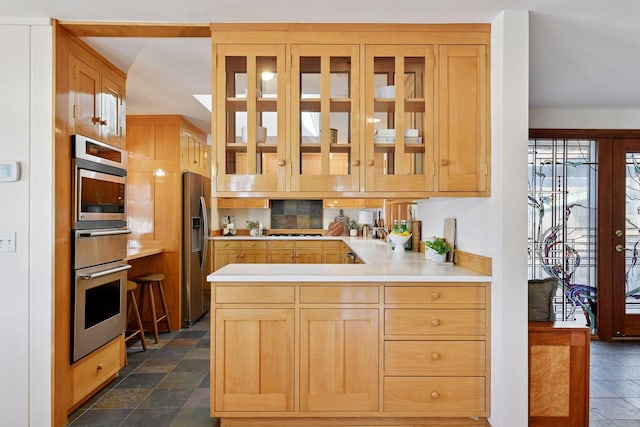 kitchen with a peninsula, light countertops, appliances with stainless steel finishes, stone finish floor, and glass insert cabinets