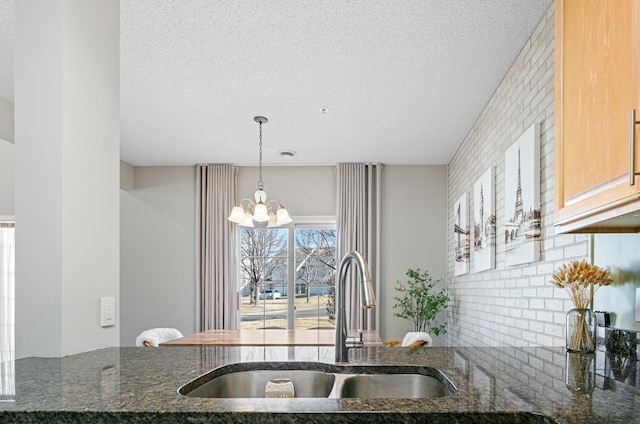 kitchen with a sink, a textured ceiling, an inviting chandelier, and dark stone countertops