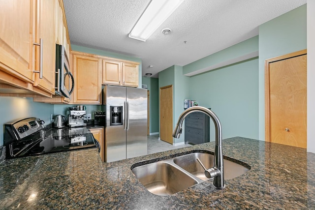 kitchen with light brown cabinetry, dark stone counters, appliances with stainless steel finishes, a textured ceiling, and a sink