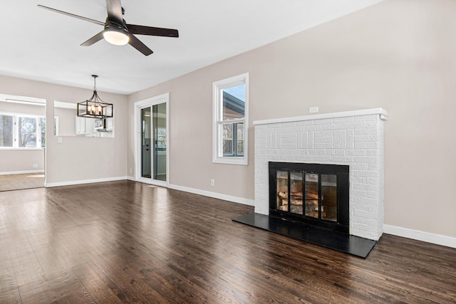 unfurnished living room with ceiling fan with notable chandelier, a brick fireplace, wood finished floors, and baseboards