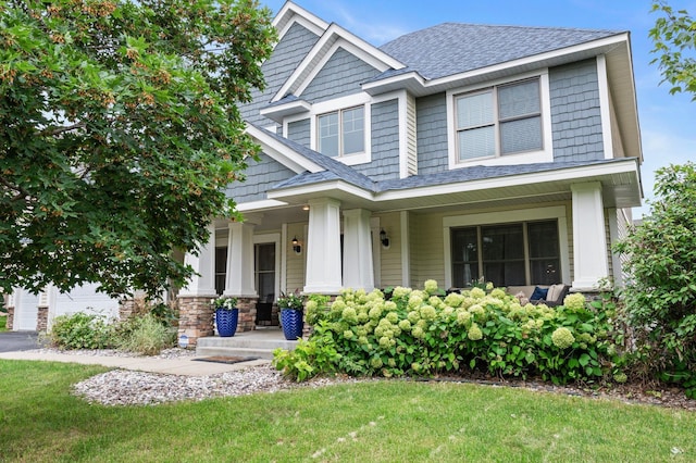 craftsman house featuring covered porch, a shingled roof, stone siding, and a front lawn