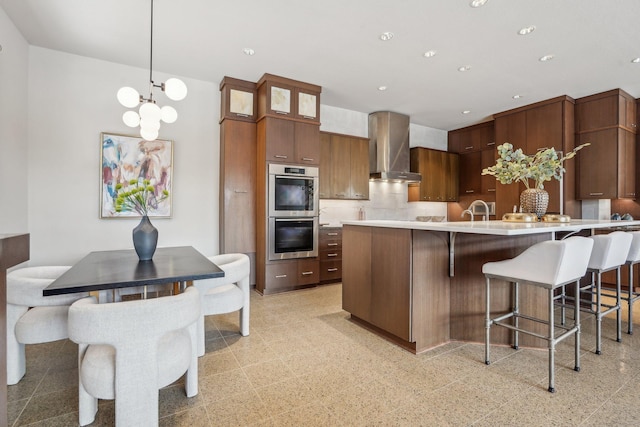 kitchen with recessed lighting, light countertops, double oven, wall chimney range hood, and a kitchen bar