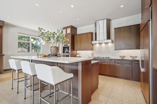 kitchen with a center island with sink, baseboards, decorative backsplash, wall chimney range hood, and recessed lighting