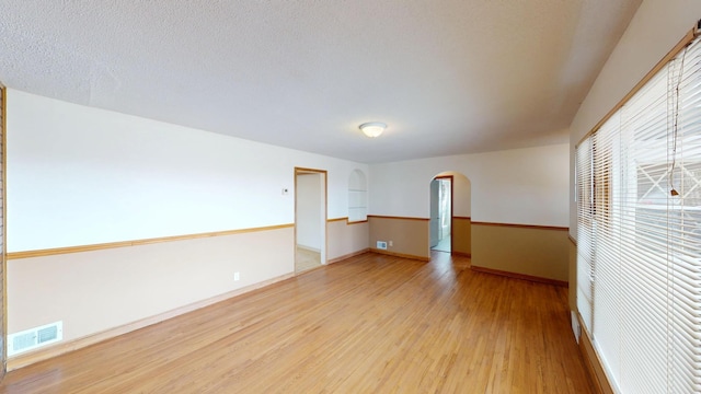 empty room featuring arched walkways, visible vents, light wood-style floors, a textured ceiling, and baseboards