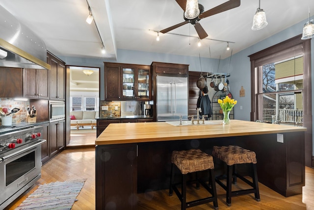kitchen with wall chimney exhaust hood, premium appliances, a ceiling fan, wood counters, and rail lighting