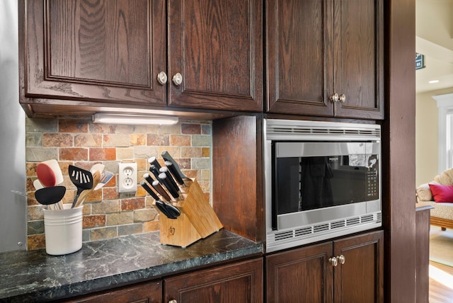 kitchen with stainless steel microwave, dark brown cabinets, decorative backsplash, and dark stone countertops
