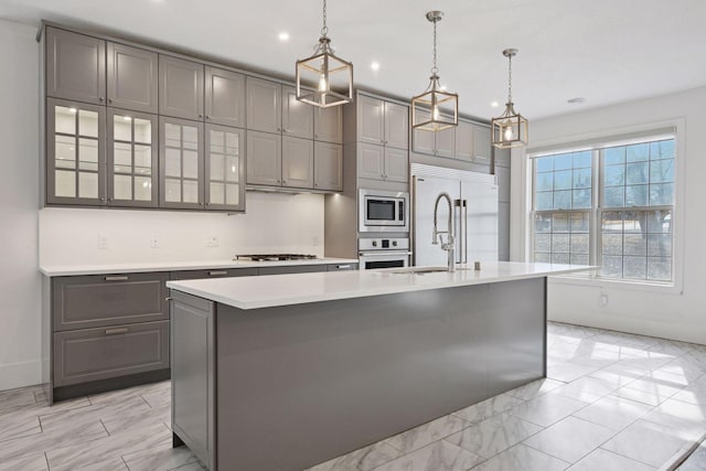 kitchen featuring a center island with sink, glass insert cabinets, built in appliances, light countertops, and gray cabinetry