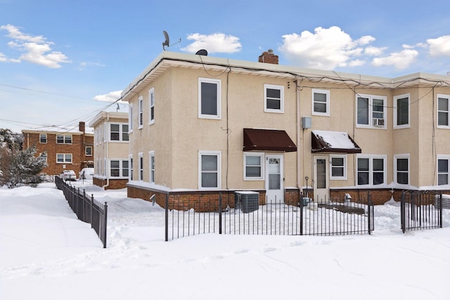 snow covered property with central AC unit, fence, a chimney, and stucco siding