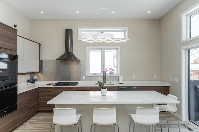 kitchen featuring black appliances, wall chimney range hood, modern cabinets, and a kitchen breakfast bar