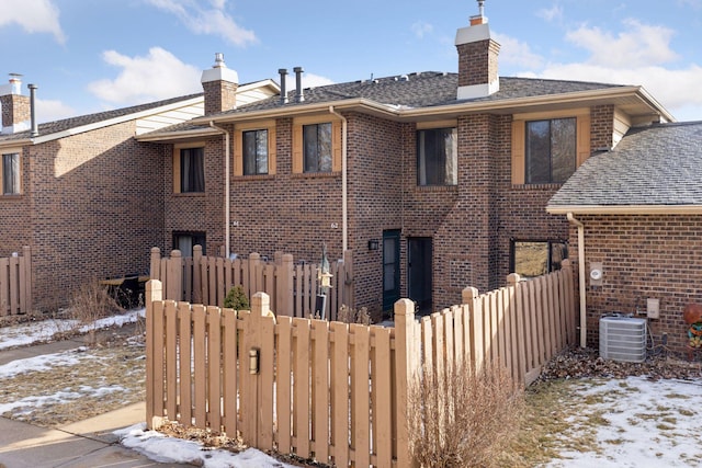 snow covered house featuring central AC, brick siding, a chimney, and fence