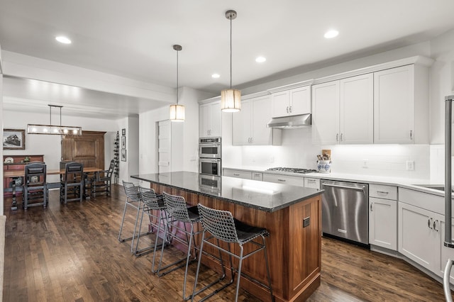 kitchen with under cabinet range hood, appliances with stainless steel finishes, dark wood finished floors, and a kitchen breakfast bar