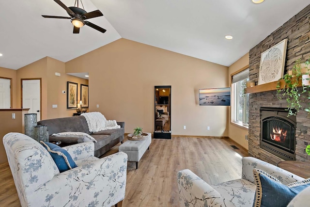 living area with light wood-type flooring, lofted ceiling, a ceiling fan, a stone fireplace, and baseboards