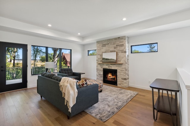 living room featuring light wood finished floors, baseboards, a tray ceiling, a stone fireplace, and recessed lighting