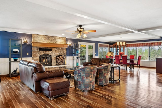 living room featuring hardwood / wood-style flooring, ceiling fan with notable chandelier, beam ceiling, wainscoting, and a stone fireplace