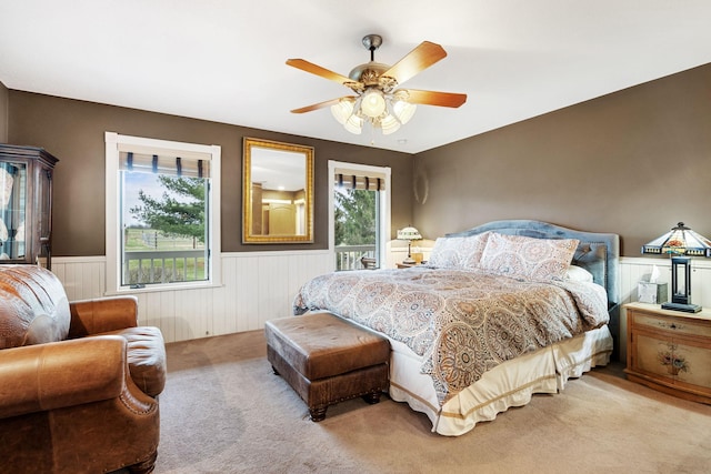 bedroom featuring a ceiling fan, multiple windows, light carpet, and a wainscoted wall