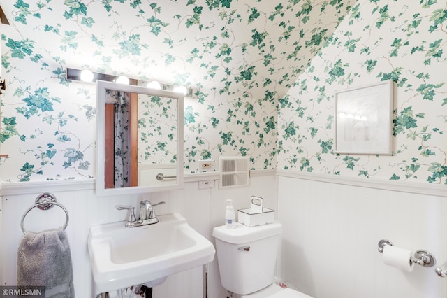 bathroom with toilet, wallpapered walls, a sink, and wainscoting